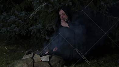 A mysterious hooded monk prays by the fire with a rosary.