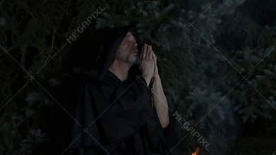 A hooded ascetic monk prays by a fire deep in the forest.