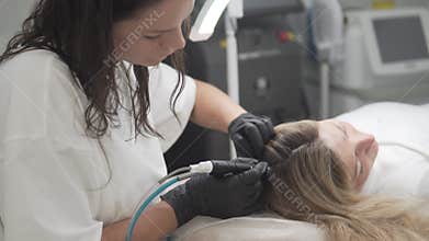 Female cosmetologist carefully performs HydraFacial Keravive scalp treatment on a blonde client in a professional