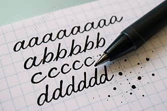 Black ink pen writing cursive lowercase letters on grid notebook paper with ink splatters and close up detail of calligraphy