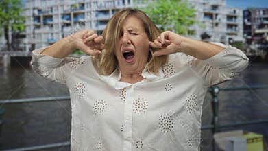 Middle aged caucasian woman covering ears with fingers against canal water by street railing; noise irritation