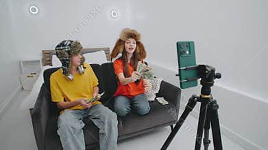 Two young influencers funny fluffy hats with cash throwing in a cozy room while filming content for their social media