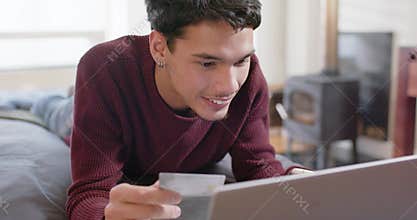African American adult male seeing payment prompt with card, paying on silver laptop on bed