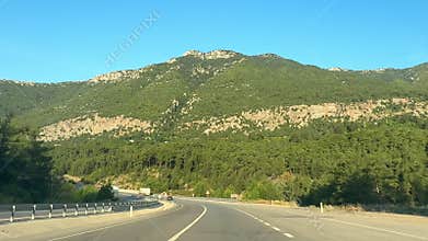 A scenic view of a highway in Turkiye, surrounded by lush green mountains under a clear blue sky. The road curves gently through