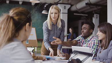 Blonde woman team leader giving direction to mixed race team of young guys. Creative business meeting at modern office.