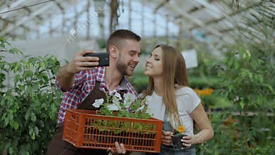 Cheerful loving couple gardeners taking selfie picture on smartphone camera and kissing while working in greenhouse