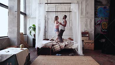 Multiethnic couple in pajamas having fun together. African man and european woman dancing, jumping on bed, laughing.