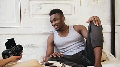 Handsome african model man in pajamas posing to photographer. Woman taking photos on old moving-film camera.