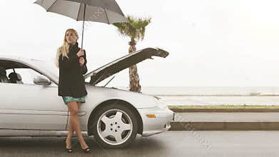 A young blonde girl stands beside her broken car under the umbrella