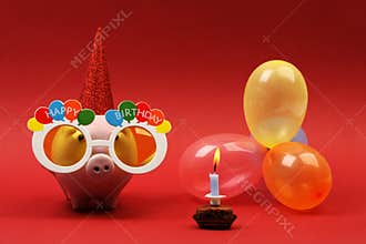 Piggy bank with sunglasses Happy birthday, party hat and multicolored party balloons on red background