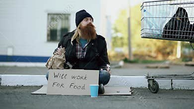 Young man helps to homeless person and giving him some money while beggar drink alcohol and sit near shopping cart at