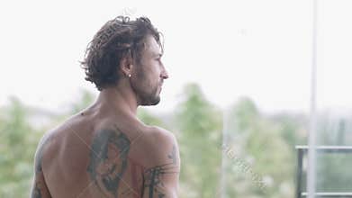Handsome muscular man with tattoos on his bare back step out onto the open balcony