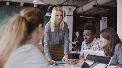 Blonde woman team leader giving direction to mixed race team of young guys. Creative business meeting at modern office.