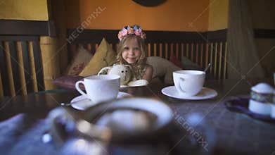 A little happy girl is sitting on the couch at a cafe and hugging her stuffed rabbit.