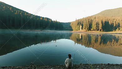 Handsome young thoughtful man in embroidery shirt throwing stones into the water and looking on the picturesque mountain