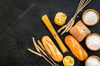 Farinaceous food. Fresh bread and raw pasta near flour in bowl and wheat ears on black background top view copy space