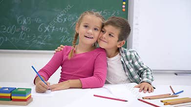 School love, classmates write during lesson at table and then look at camera and smile on background of blackboard