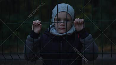 Lonely unhappy, miserable young boy behind and clinging to fence