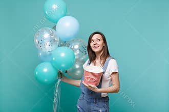 Pensive young woman in denim clothes looking up holding bucket of popcorn celebrating with colorful air balloons