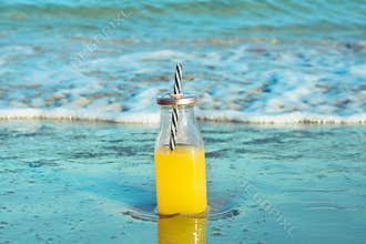 Glass bottle with fresh tropical fruits juice with straw standing on beach sand shore blue turquoise foamy sea waves