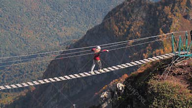 People in helmets walking along hanging rope bridge over abyss