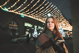 Portrait of a cute lady in warm clothing uses a smartphone,looks in the direction and smiles at an evening walk