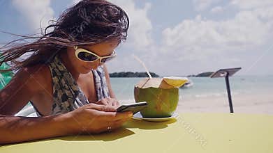 Relaxing young brunette woman wearing sunglasses using cellphone and drinking a cocktail from a coconut in the beach