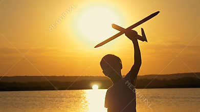 Happy child on a background of an orange sky and lake in summer at sunset, playing with a toy airplane.