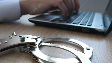 Man working on a laptop and handcuffs on a desk. Online cyber crime.