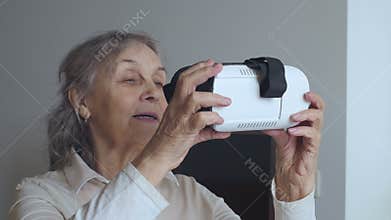 Medium Shot of Senior Woman Examines VR Glasses. Good-looking Senior Woman In White Inspect VR 360 Glasses At Home.