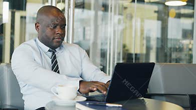 African American businessman thinking, printing and working on his laptop in glassy cafe during lunch break. He is