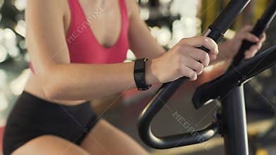 Healthy slim female wearing fitness bracelet riding exercise bike at sports club