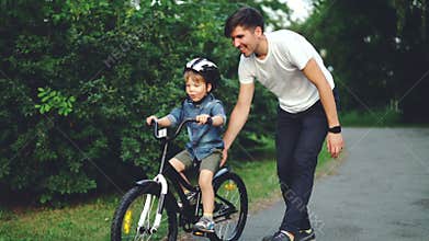 Slow motion of excited boy riding bicycle and laughing while his careful father is helping him holding bike and teaching
