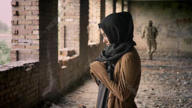 Young muslim woman in hijab standing in abandoned building, soldier walking in background, military