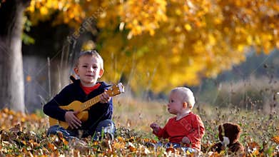 Portrait of amusing child sing at guitar to baby brother in autumn park