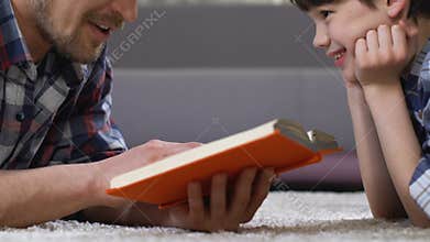 Father reading fairytale to son lying on floor, home leisure preschool education