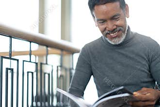 Portrait of attractive mature asian man retired with stylish short beard sitting, smiling and reading magazine books