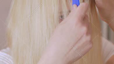 A blond woman with long hair is combing in front of a mirror, preparing her head for coloring hair roots.