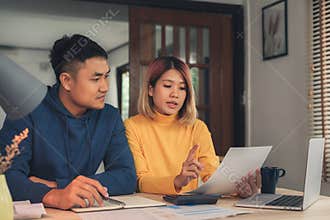 Young asian couple managing finances, reviewing their bank accounts using laptop computer and calculator at modern home.