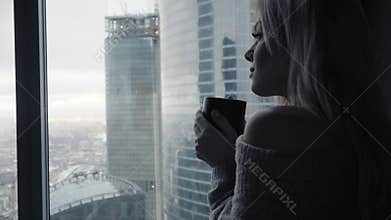 Beautiful blonde woman holding ceramic cup of tea or coffee enjoy. Tea time in bed room. Looking at window and drink tea
