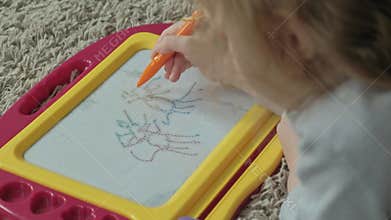A little girl with red wavy hair lies on the floor and draws on a magnetic board. The concept of the educational process