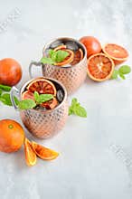 Blood orange Moscow mule alcohol cocktail with fresh mint leaves and ice in copper mugs on a gray concrete background.