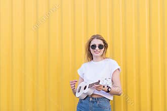 Attractive, stylish girl stands on the background of a yellow wall with ukulele in her hands and looks at the camera