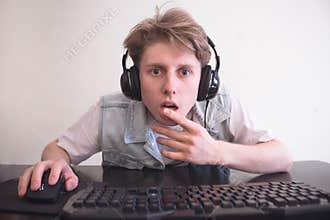 Surprised teenager in the headphones sits at home in the room behind and plays video games on a computer
