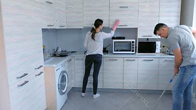Husband and wife are cleaning in the kitchen together. Man washes the floor with a mop and woman wipes the furniture.