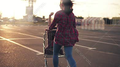 Back view of a young woman pushing the grocery cart, while her friend is sitting inside during sunset. Lens flare