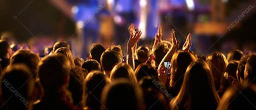 Audience with hands raised at a music festival and lights streaming down from above the stage.