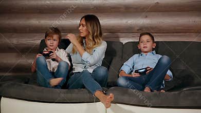 Mother and two sons sitting on the sofa in his house playing video games with wireless joystick. Happy people in the