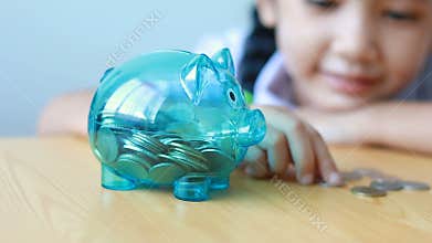 Asian little girl in Thai kindergarten student uniform putting money coin into clear piggy bank on wooden table metaphor money sav