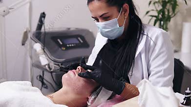 Professional cosmetologist is applying face cream and making face massage for young woman with blemish skin lying on the